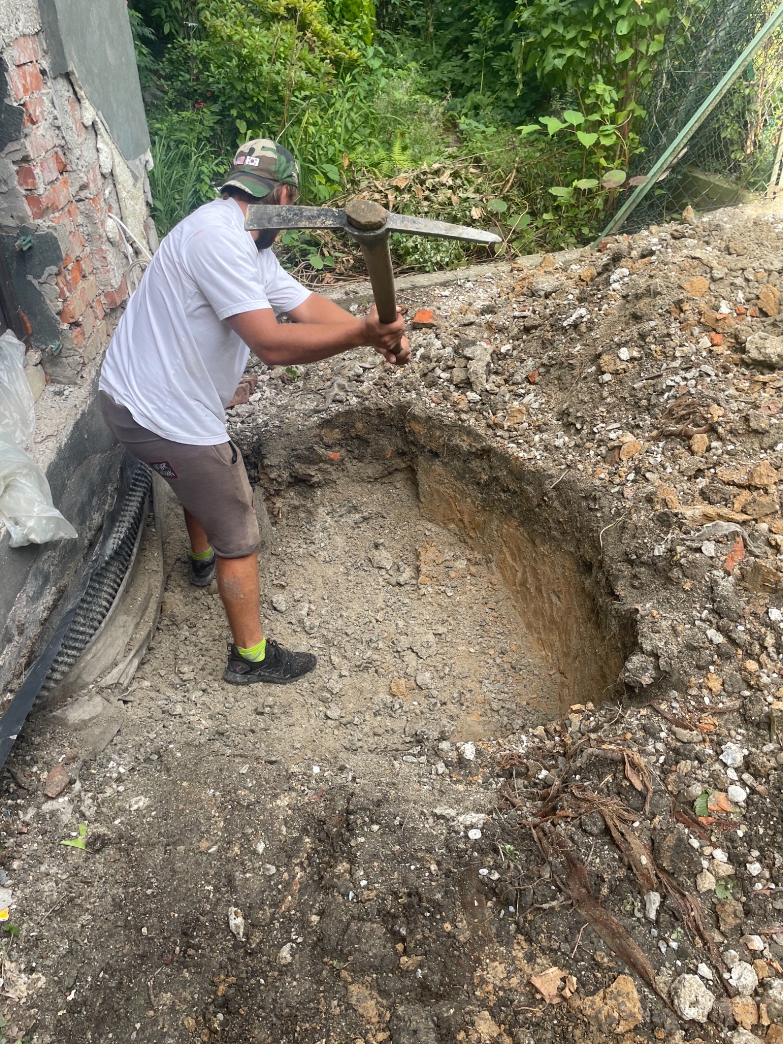 Mężczyzna w czapce z daszkiem kopie dół w ziemi kilofem przy fundamentach budynku, widoczna izolacja fundamentów.