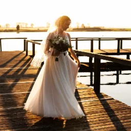 Panna młoda w zwiewnej sukni i welonie, trzymająca bukiet, spaceruje po drewnianym pomoście nad jeziorem w promieniach zachodzącego słońca.