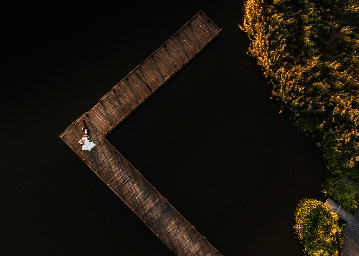 Widok z góry na parę młodą spacerującą po drewnianym pomoście nad wodą, otoczonym trzcinami w ciepłym świetle zachodzącego słońca.