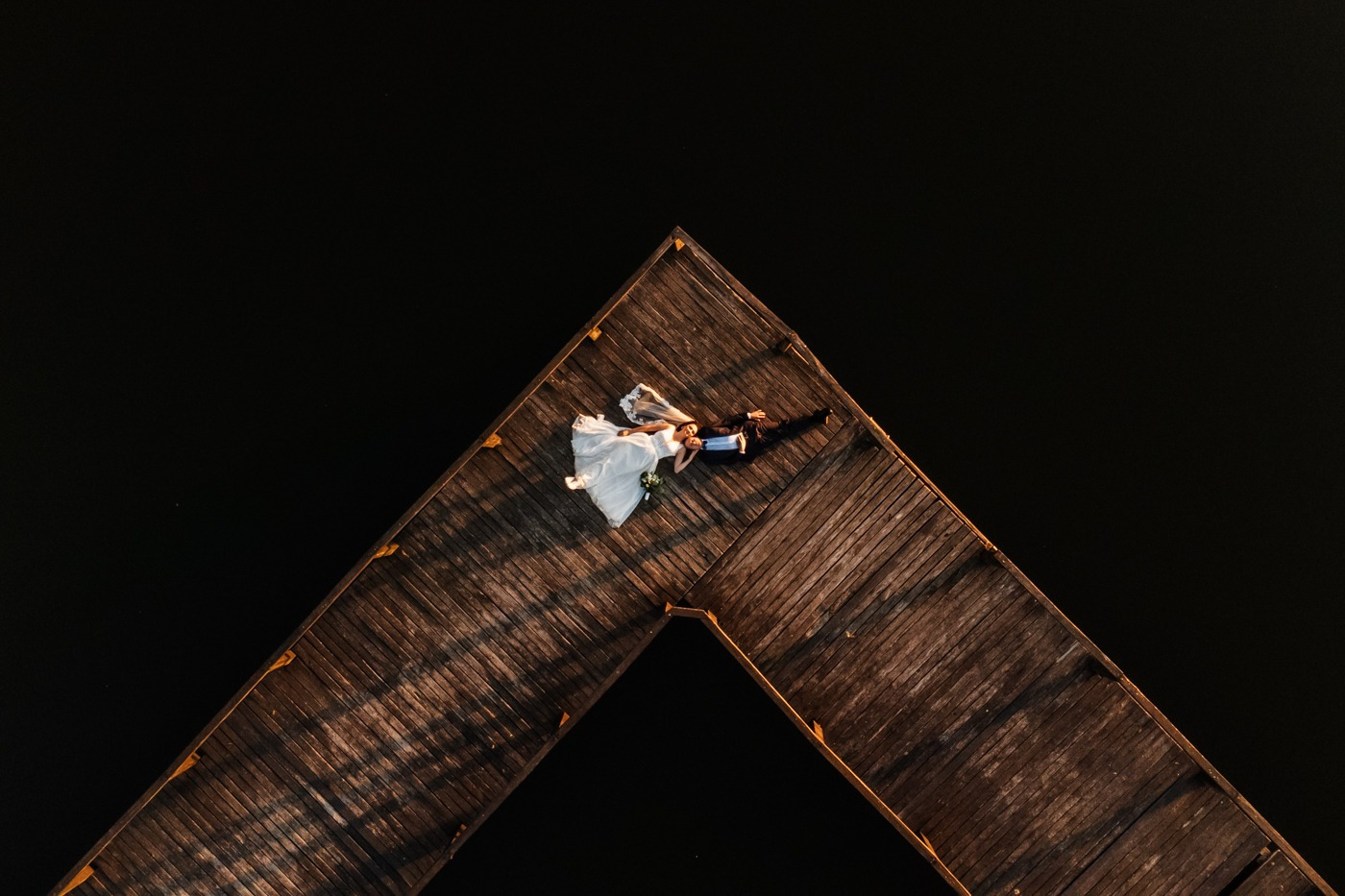 Para młoda leżąca na drewnianym pomoście w kształcie strzały, widok z lotu ptaka, ciemna woda w tle.