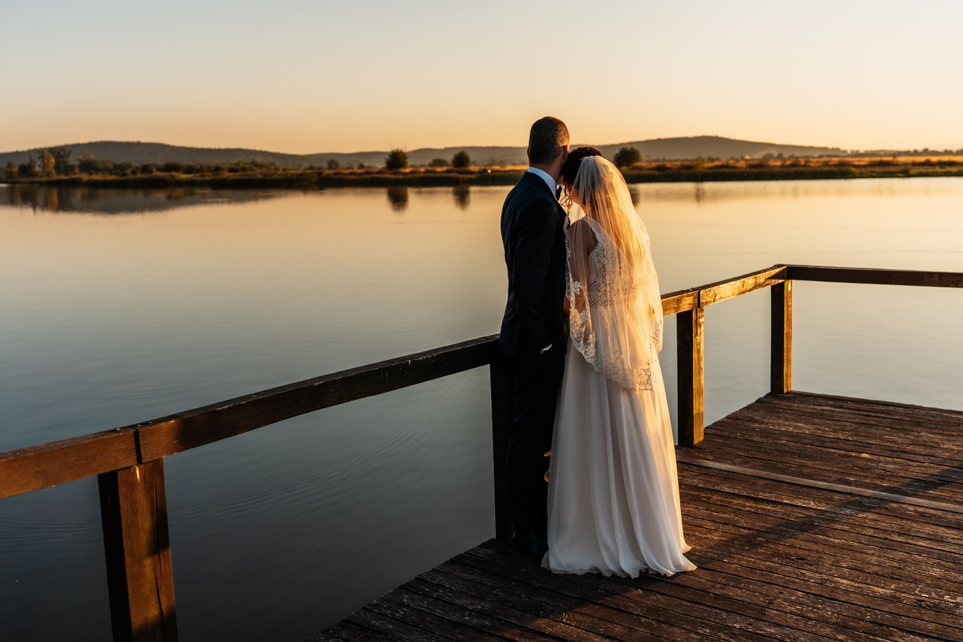 Para młoda, obejmująca się na drewnianym pomoście nad spokojnym jeziorem o zachodzie słońca, z widokiem na zalesione wzgórza w oddali.