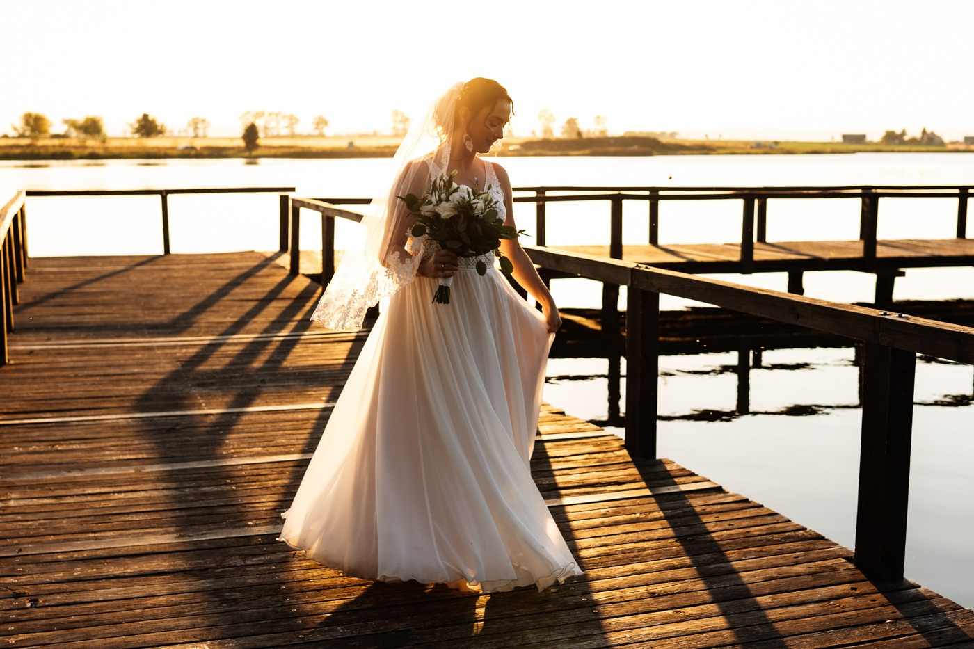 Panna młoda w zwiewnej sukni i welonie, trzymająca bukiet, spaceruje po drewnianym pomoście nad jeziorem w promieniach zachodzącego słońca.