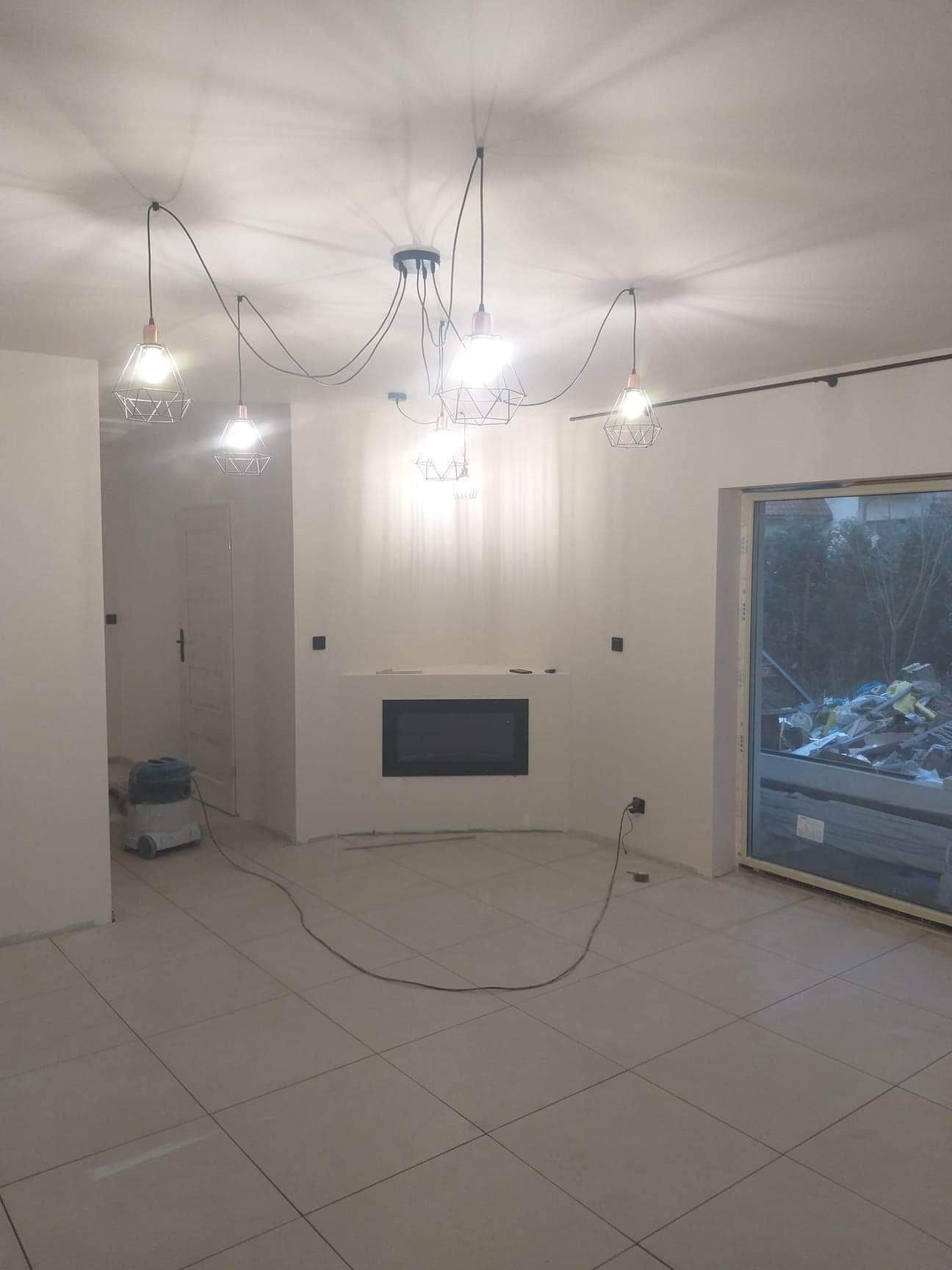 Wnętrze po remoncie z nową podłogą z płytek, świeżo pomalowanymi ścianami, nowoczesnym kominkiem i designerską lampą z kilkoma żarówkami w metalowych kloszach, widoczny odkurzacz przemysłowy.