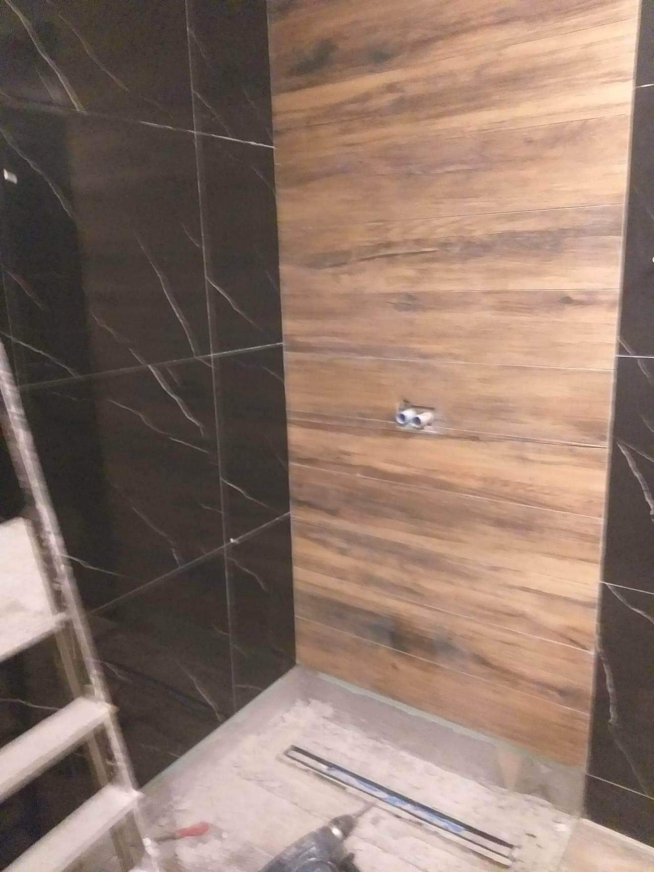 Montaż płytek imitujących drewno na ścianie łazienki, widoczne czarne płytki z marmurowym wzorem, drabina malarska i narzędzia na podłodze.