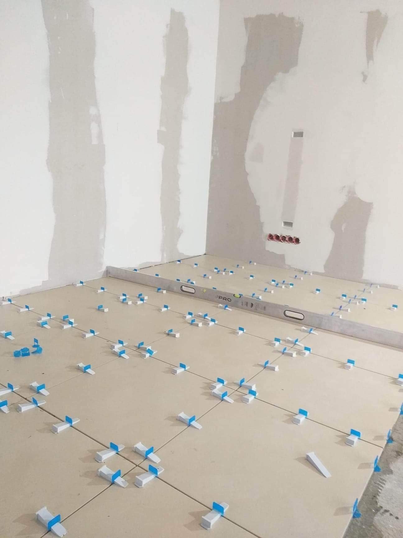 Ułożone jasne płytki podłogowe z niebieskimi krzyżykami dystansowymi, poziomica PRO na podłodze, ściany w trakcie gipsowania.