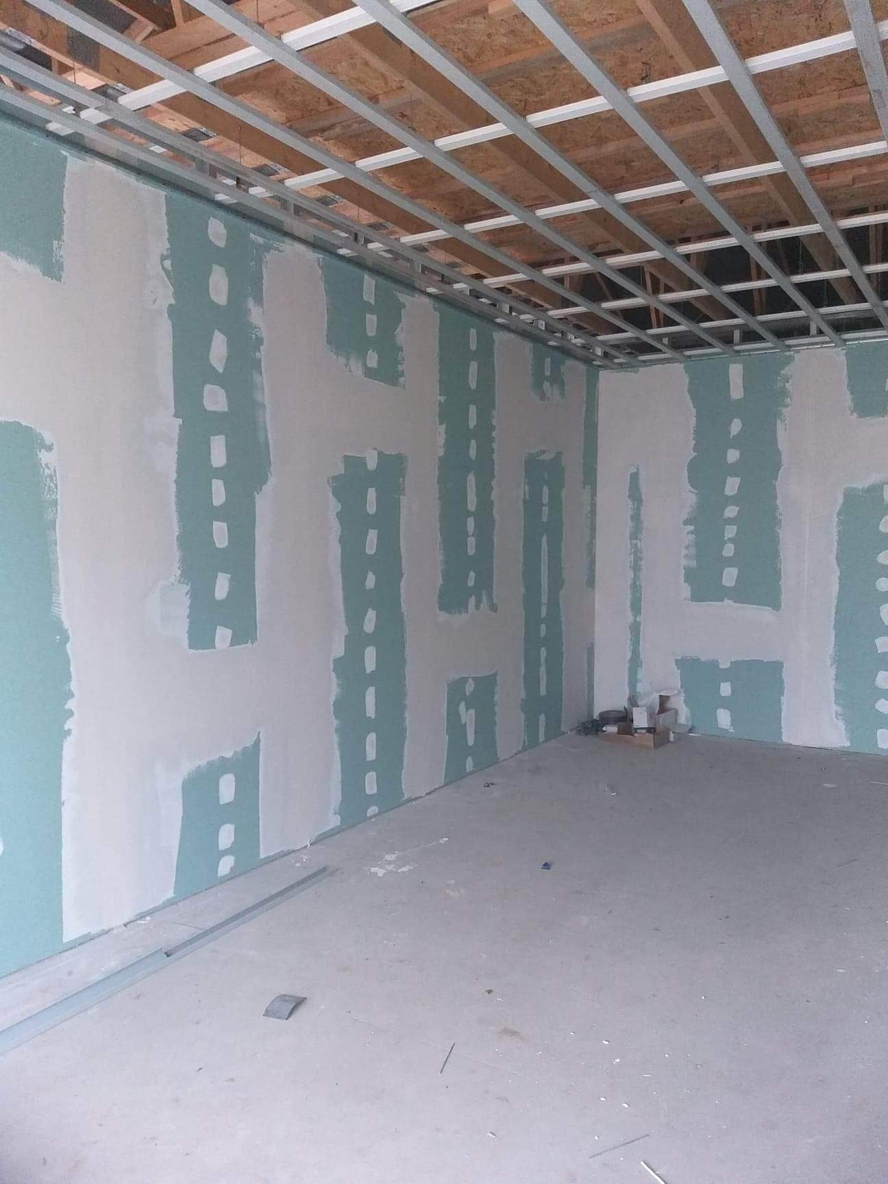 Wnętrze w trakcie prac wykończeniowych: ściany z płyt kartonowo-gipsowych z widocznymi łączeniami, metalowy stelaż sufitowy i surowy betonowy podkład podłogowy, małe pudełko z materiałami w rogu.