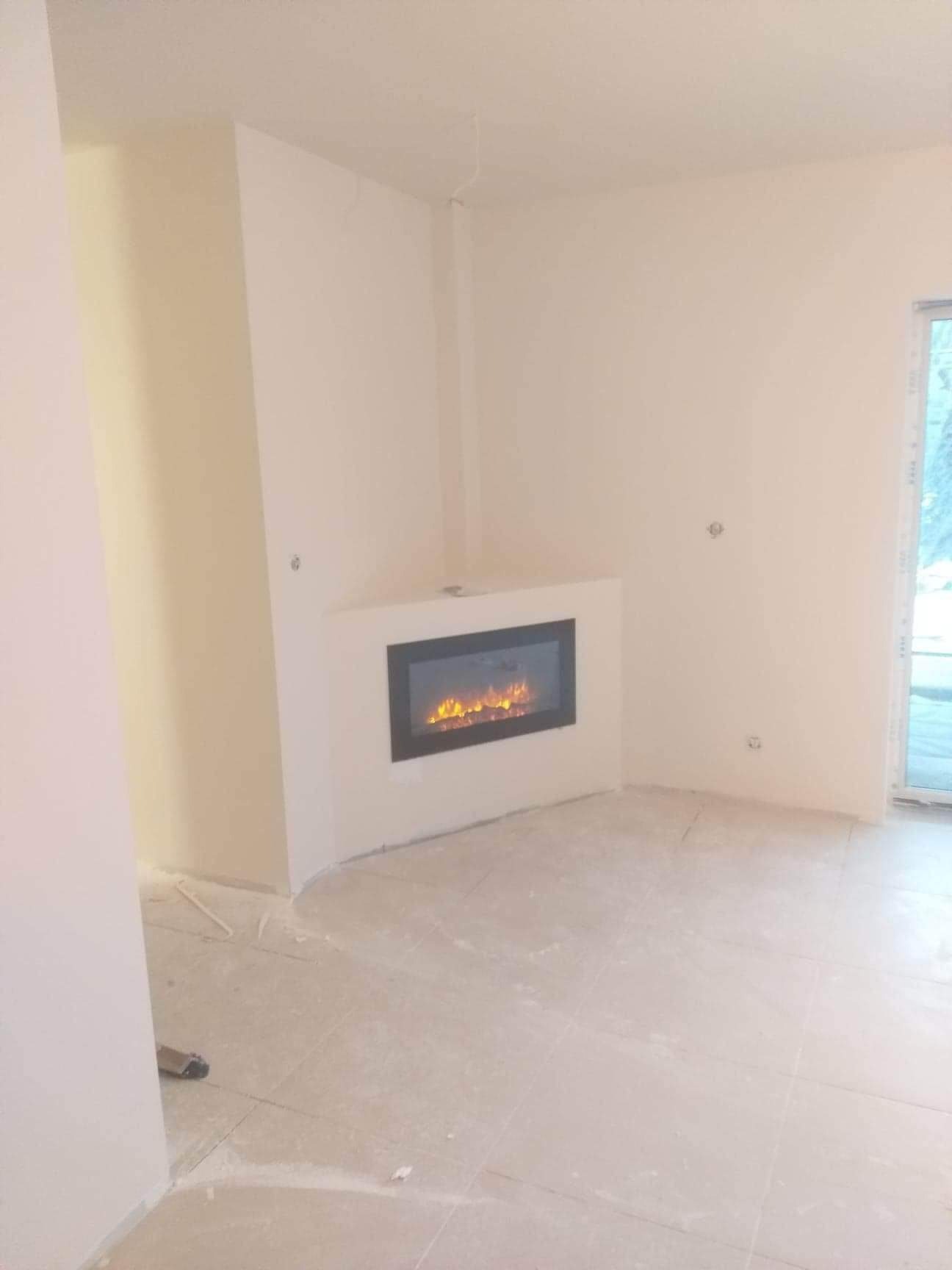 Wnętrze pokoju po remoncie z wbudowanym elektrycznym kominkiem z czarną ramą, świeżo pomalowane ściany na biało i jasna podłoga z płytek ceramicznych.