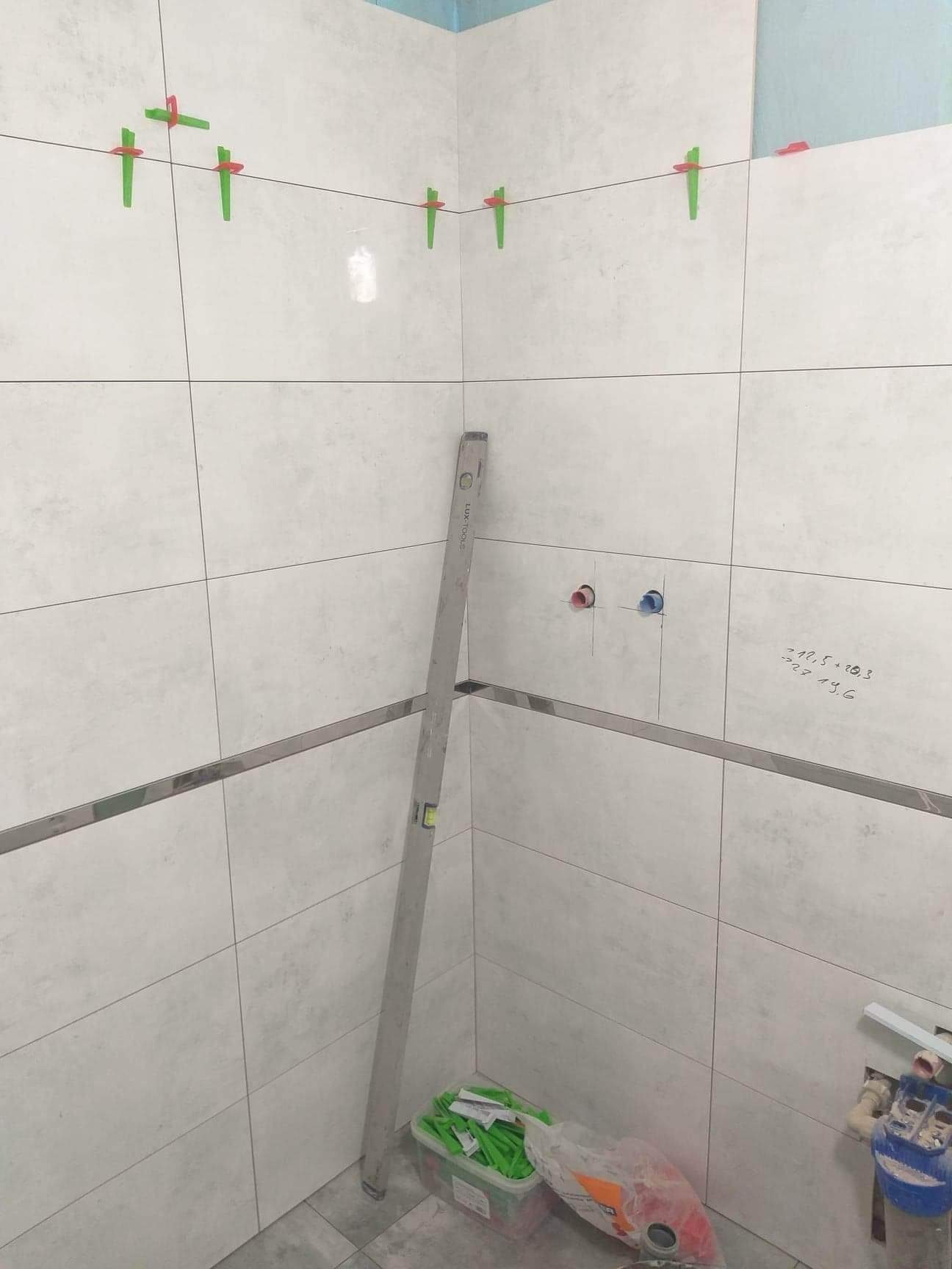 Białe płytki ceramiczne na ścianie w rogu łazienki, układane z użyciem poziomicy i zielonych krzyżyków dystansowych, widoczne przyłącza wodne i odręczne notatki na ścianie.