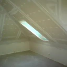 Poddasze z oknem dachowym w trakcie montażu płyt kartonowo-gipsowych, widoczne szpachlowane łączenia i wkręty, surowy beton na podłodze.