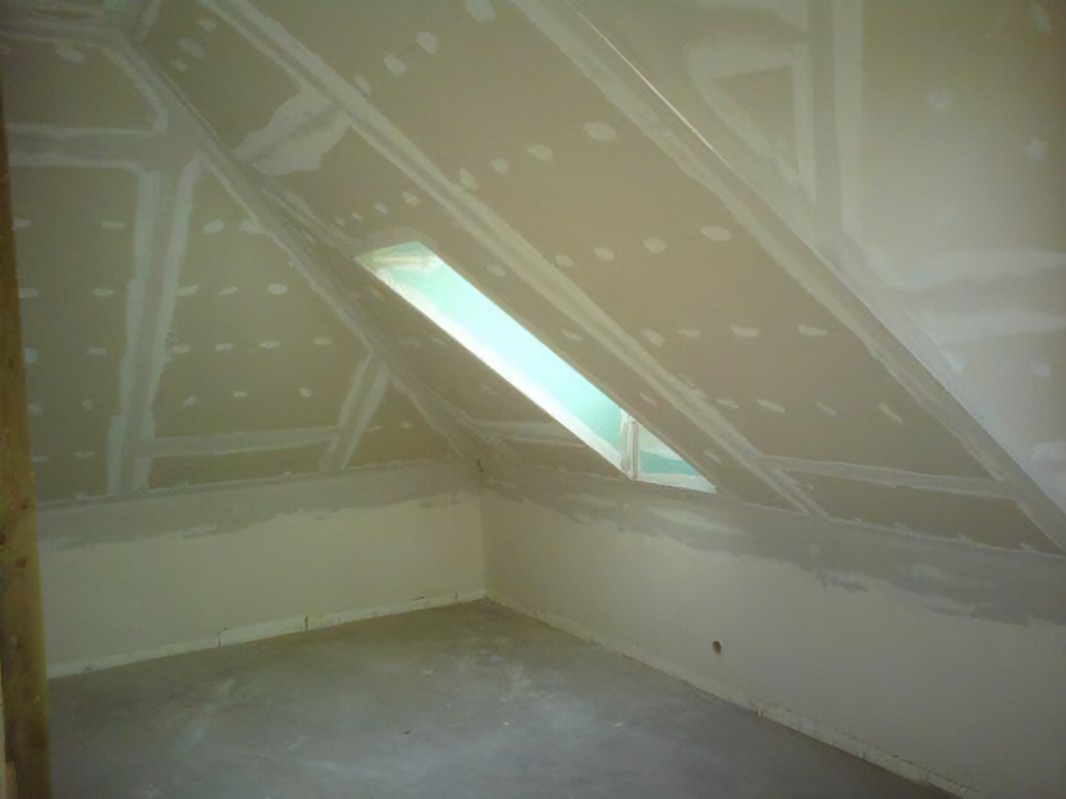 Poddasze z oknem dachowym w trakcie montażu płyt kartonowo-gipsowych, widoczne szpachlowane łączenia i wkręty, surowy beton na podłodze.