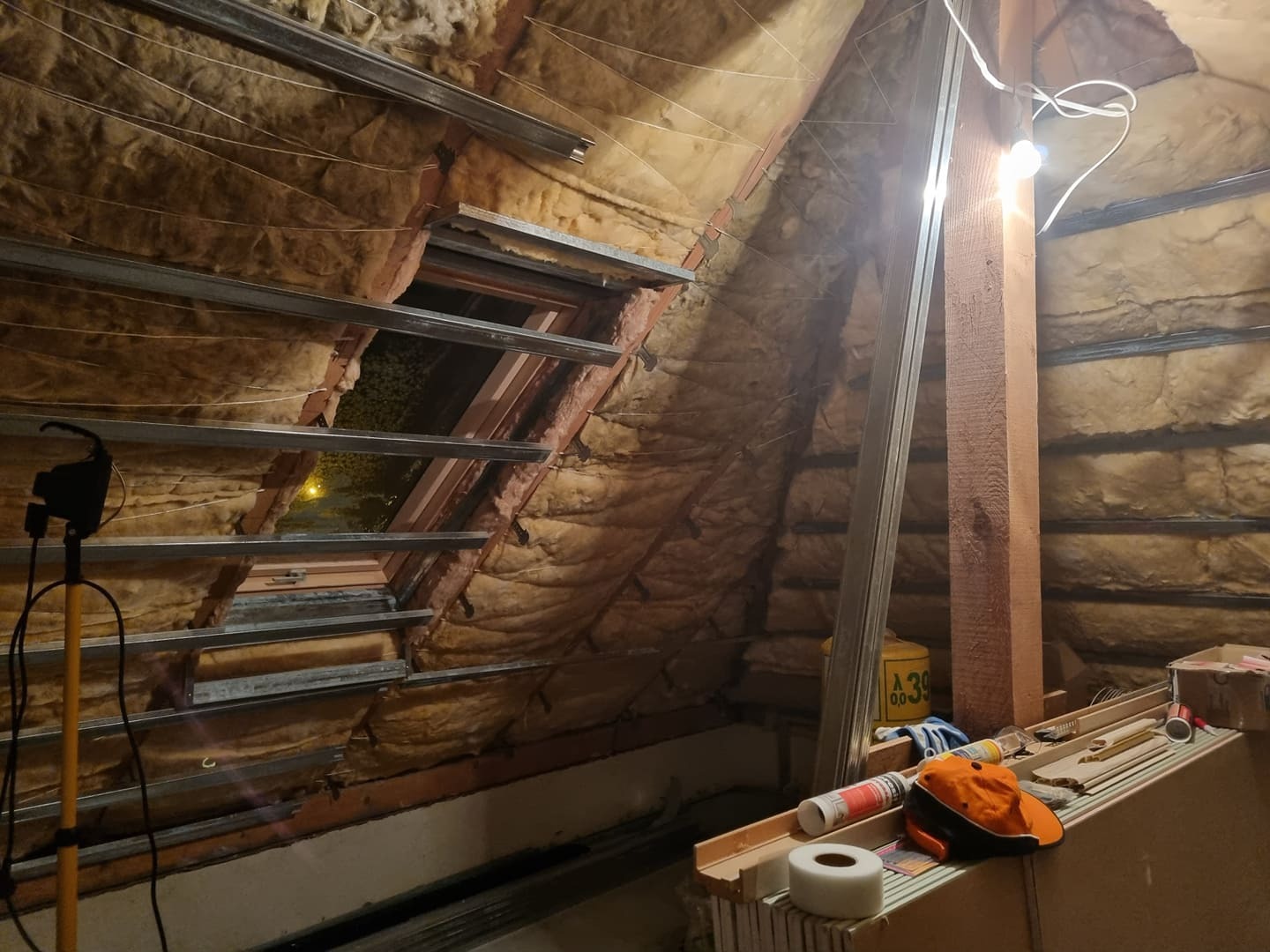 Poddasze w trakcie ocieplania wełną mineralną, widoczne metalowe profile pod płyty gipsowo-kartonowe, okno dachowe i narzędzia pracy.