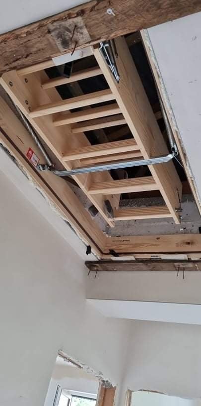 Otwór w suficie z zamontowanymi drewnianymi schodami strychowymi, widoczne sprężyny i metalowe elementy mechanizmu składania, surowe wykończenie otworu, fragment ściany z widocznymi śladami prac...
