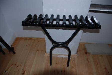 Metalowa ławeczka cmentarna z czarnymi, profilowanymi siedziskami, ustawiona na drewnianej podłodze przy białej ścianie.