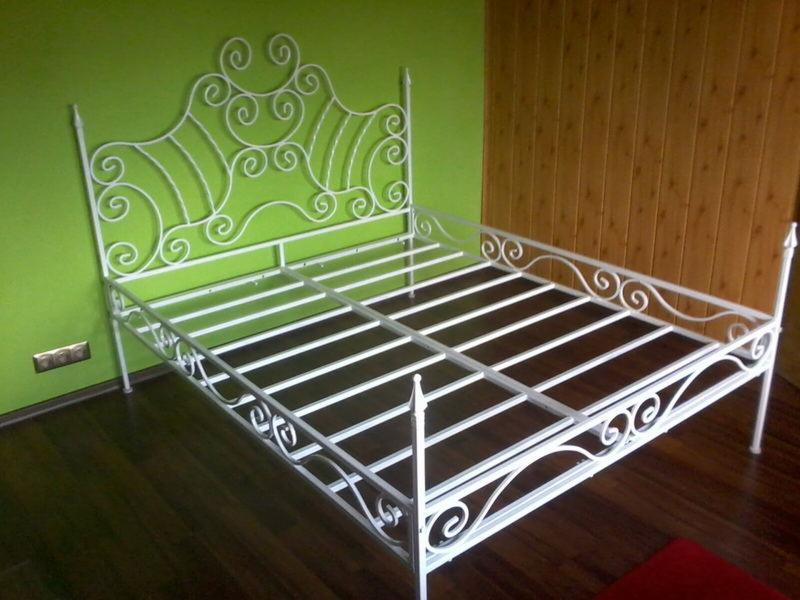 Biała, kuta rama łóżka z ozdobnymi, spiralnymi elementami na tle zielonej ściany i drewnianej boazerii.