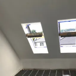 Montaż folii grzewczej na podłodze poddasza z oknami dachowymi, widoczne elementy instalacji elektrycznej i izolacji.