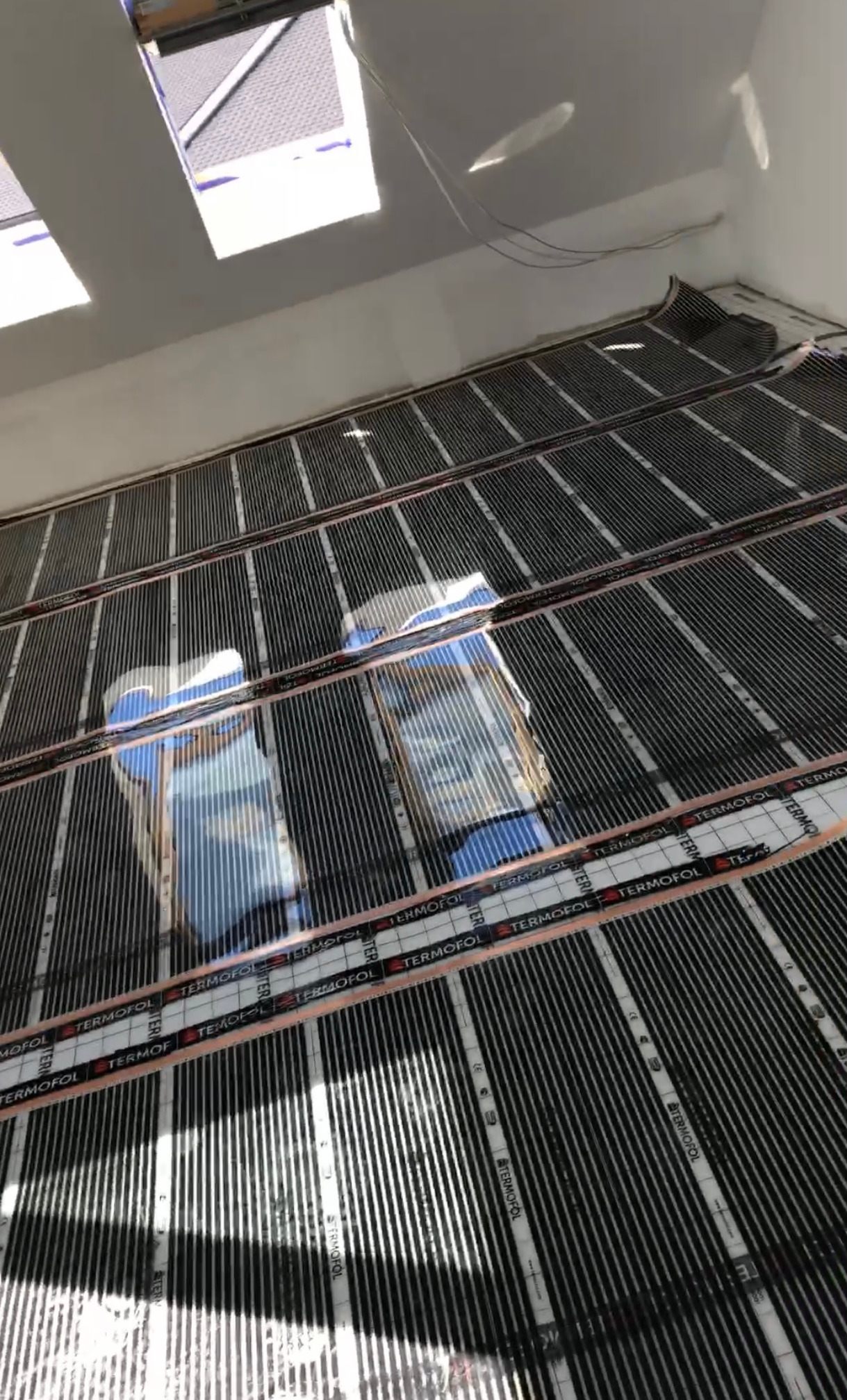 Montaż folii grzewczej Termofol na podłodze, widoczne paski folii i elementy montażowe, w tle okno dachowe.