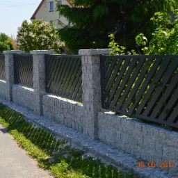 Fragment ogrodzenia z szarego kamienia i grafitowych paneli ułożonych ukośnie, widok z zewnątrz posesji w słoneczny dzień.