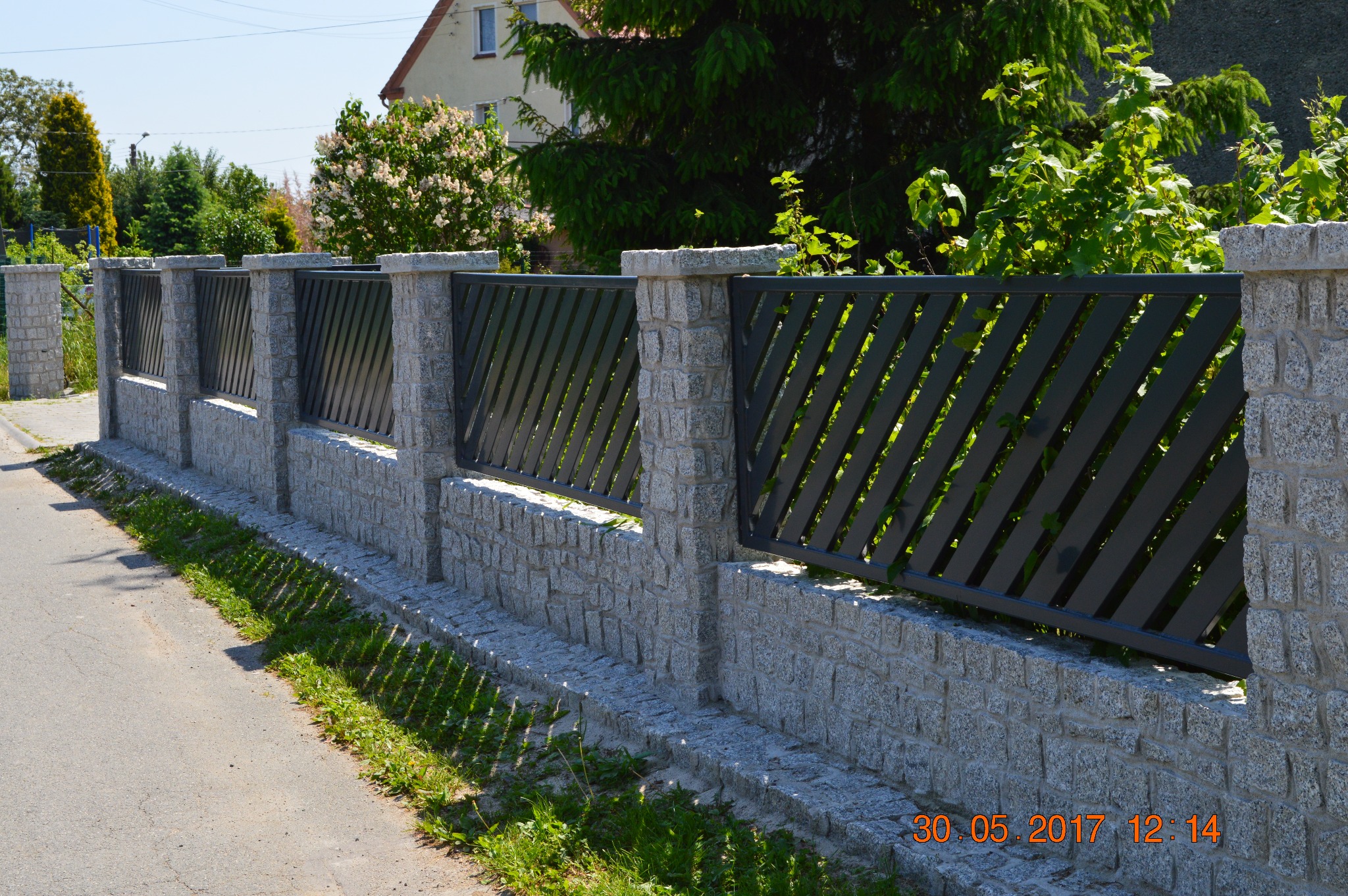 Fragment ogrodzenia z szarego kamienia i grafitowych paneli ułożonych ukośnie, widok z zewnątrz posesji w słoneczny dzień.