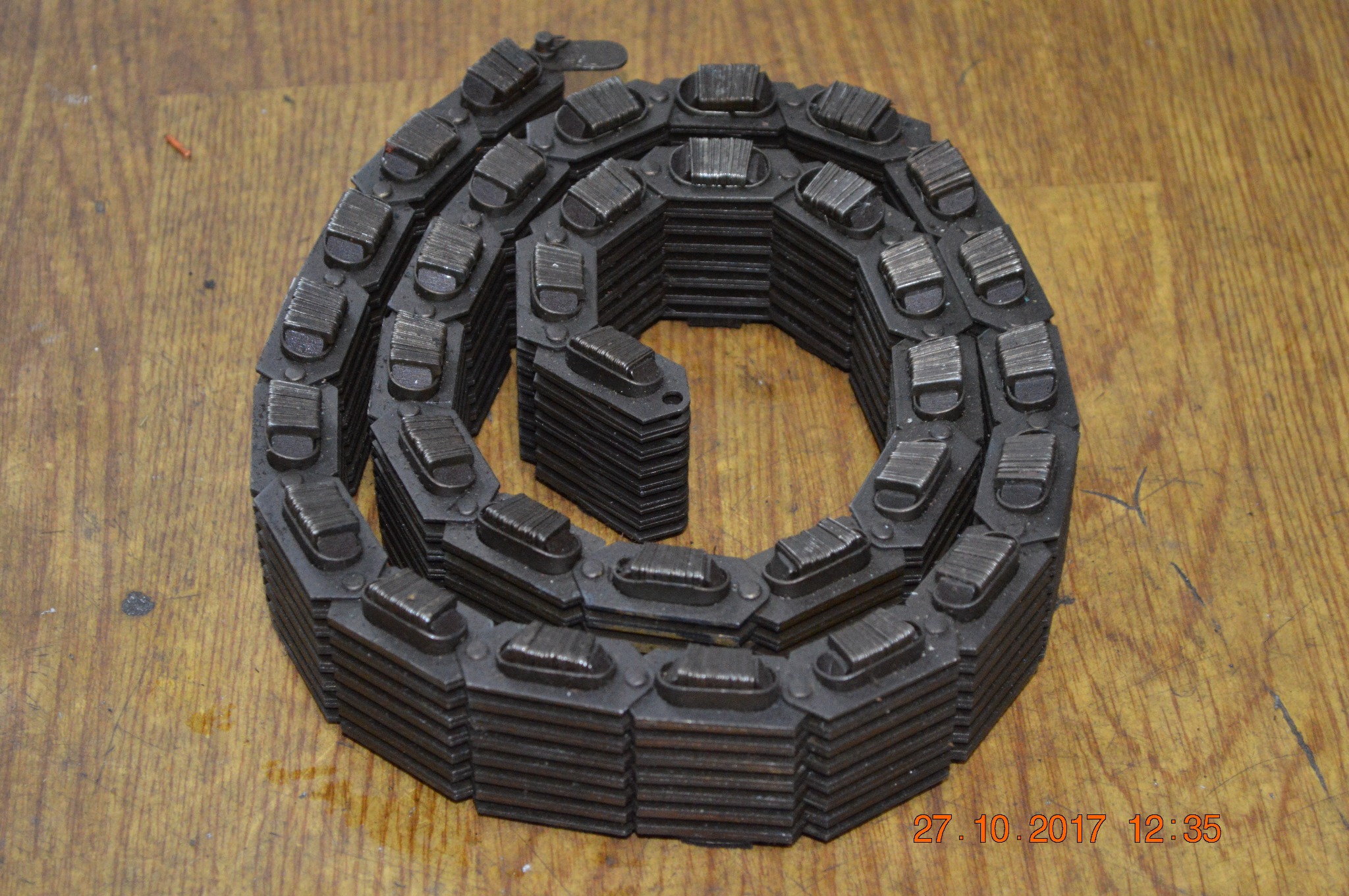 Stos metalowych ogniw łańcucha gąsienicowego ułożonych spiralnie na drewnianej powierzchni, widoczne ślady użytkowania.