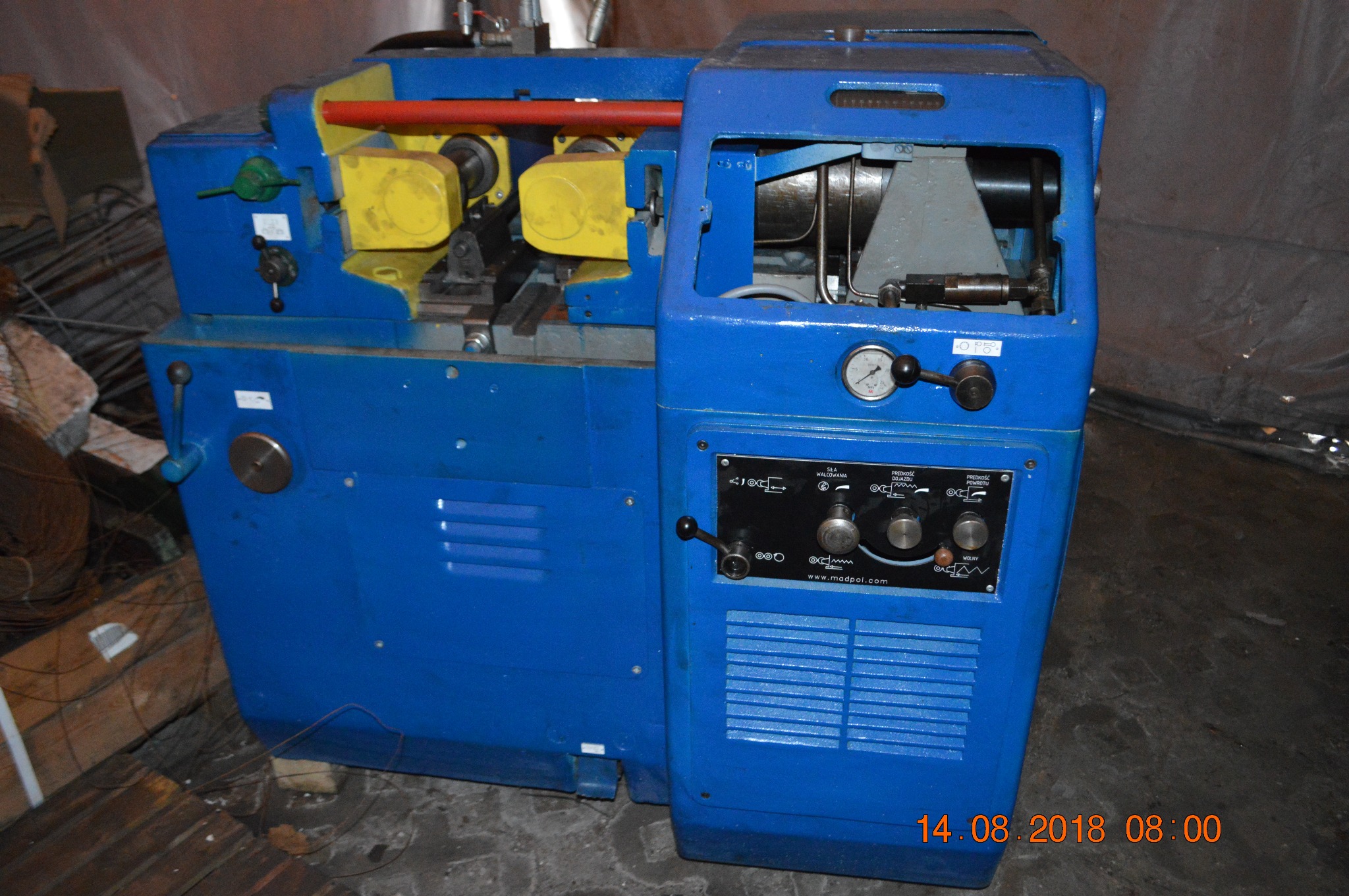 Niebieska maszyna przemysłowa z żółtymi elementami, widoczne pokrętła, wskaźniki i tabliczka znamionowa z napisem www.madpol.com, w tle drewniane elementy i przewody.