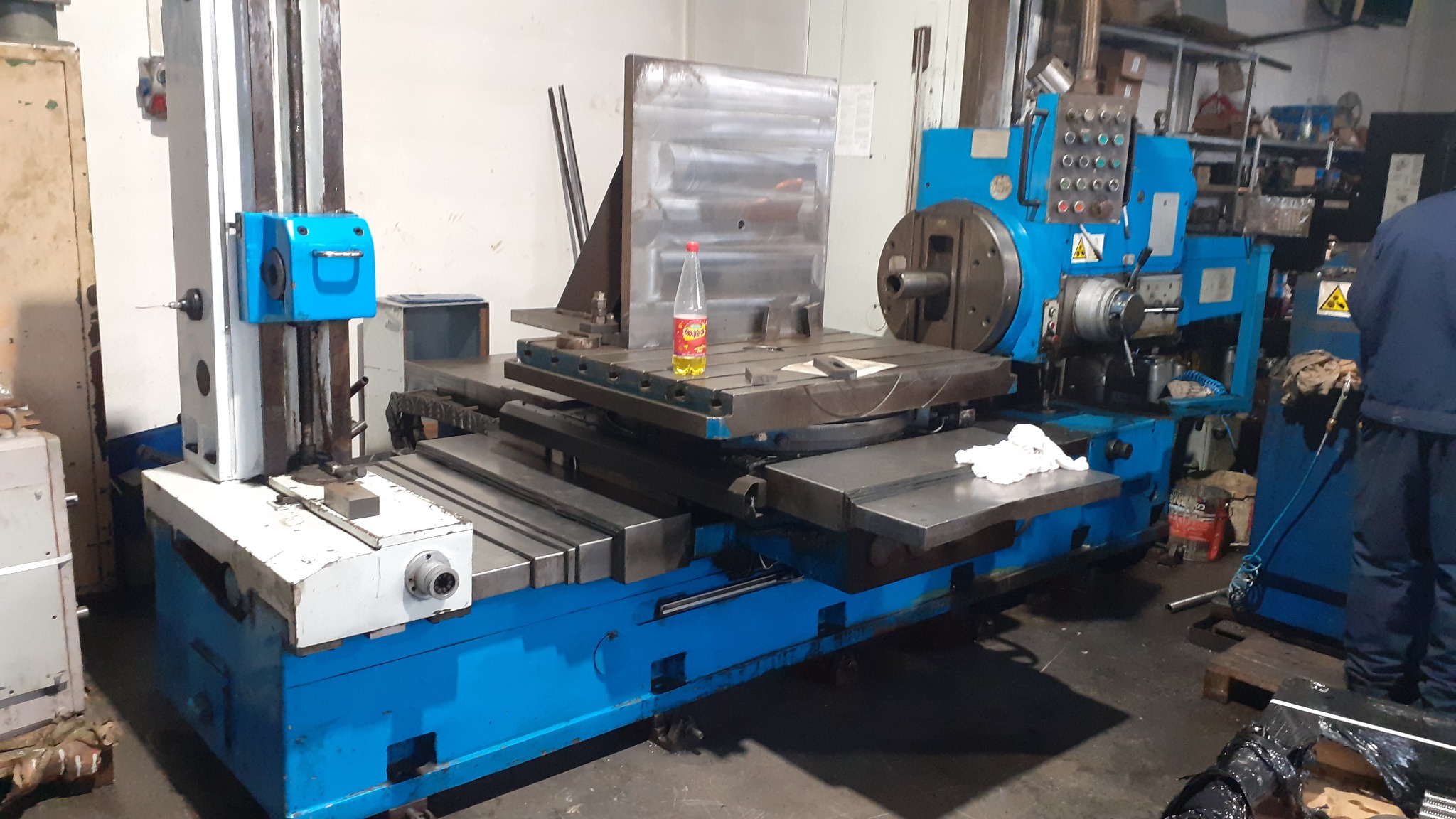 Duża, niebieska frezarka w hali produkcyjnej, z butelką oleju na stole roboczym i widoczną częścią operatora w tle.