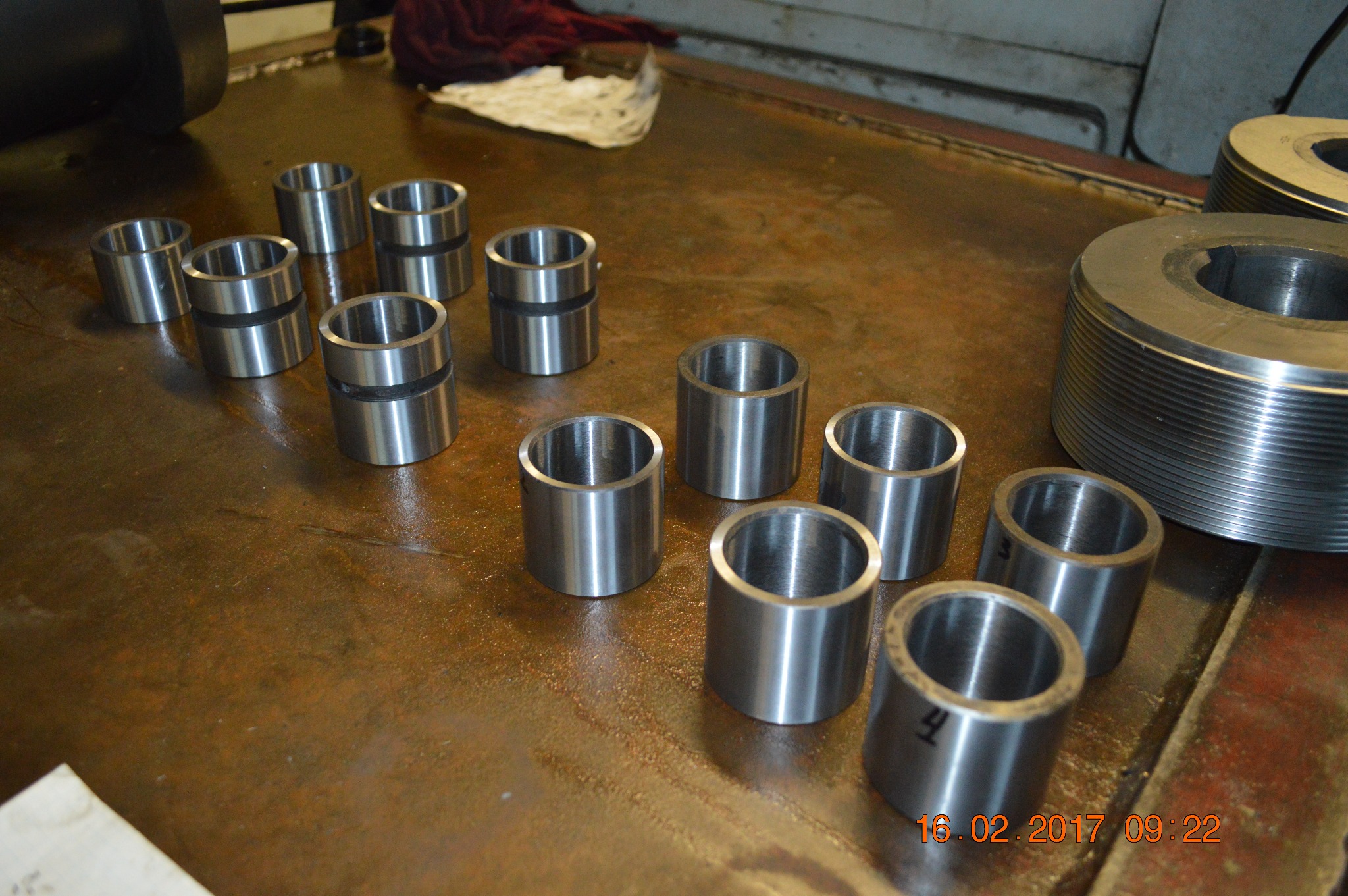 Metalowe tuleje o różnych rozmiarach, niektóre z rowkiem, ułożone na metalowym stole warsztatowym, obok większego elementu z gwintem.