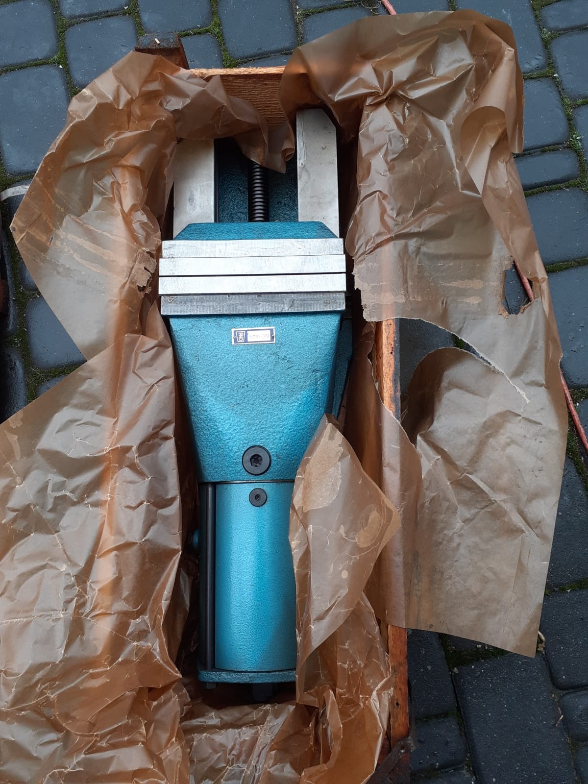 Nowa, turkusowa ślusarska imadło w drewnianej skrzyni, zabezpieczona brązowym papierem pakowym. Widoczny mechanizm śrubowy i metalowe szczęki.