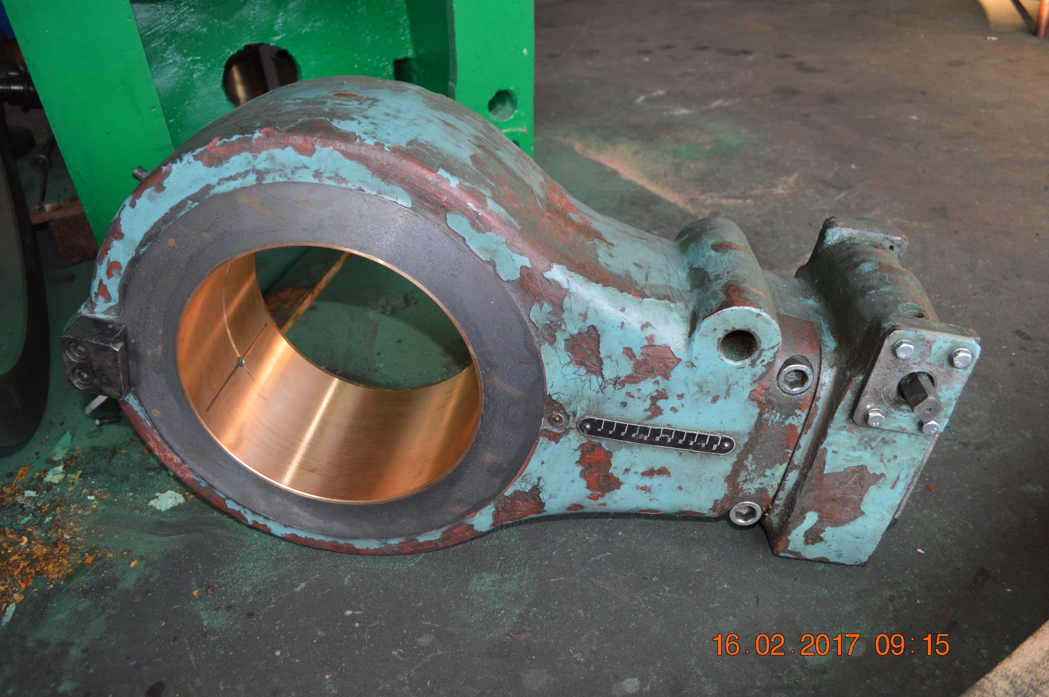 Element maszyny z brązową tuleją, pokryty warstwami farby w odcieniach zieleni i czerwieni, z widocznymi śladami użytkowania i korozji, leżący na brudnej, zielonej powierzchni.