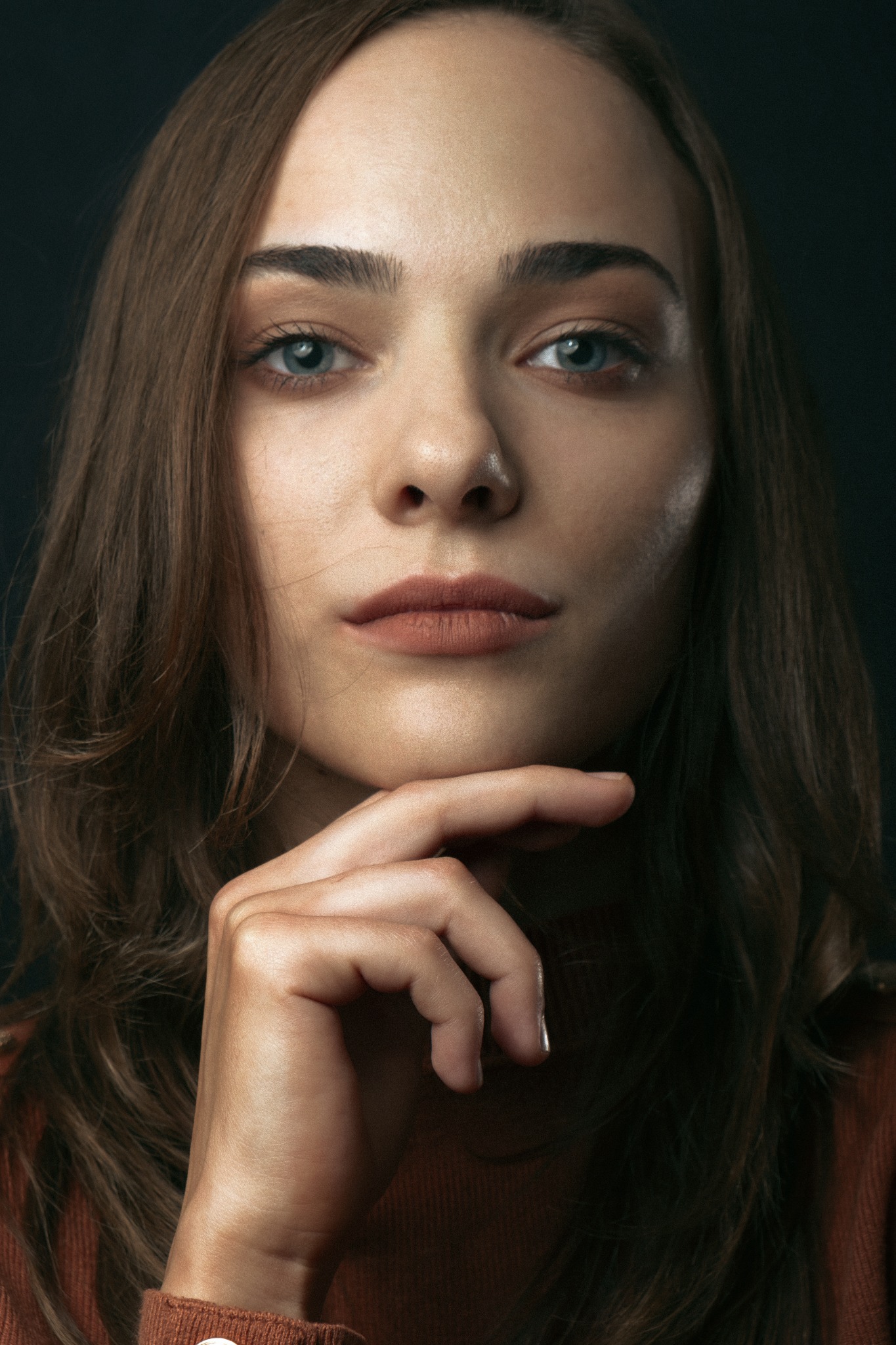 Portret kobiety z niebieskimi oczami, z ręką pod brodą, w brązowym swetrze na ciemnym tle.