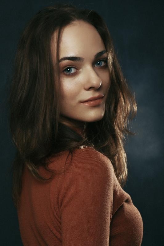 Portret młodej kobiety z długimi brązowymi włosami i niebieskimi oczami, spoglądającej przez ramię, ubrana w pomarańczowy golf na ciemnym tle.