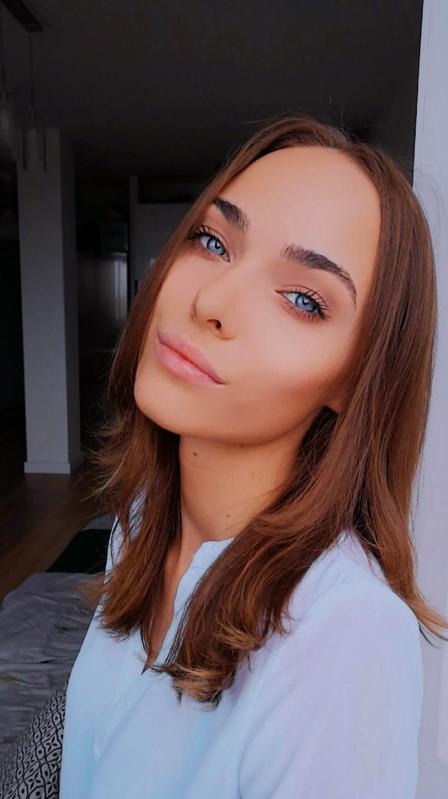 Portret młodej kobiety z brązowymi włosami i intensywnie niebieskimi oczami, lekki uśmiech, w jasnej koszuli, z delikatnym makijażem w neutralnych tonach, na rozmytym tle wnętrza.