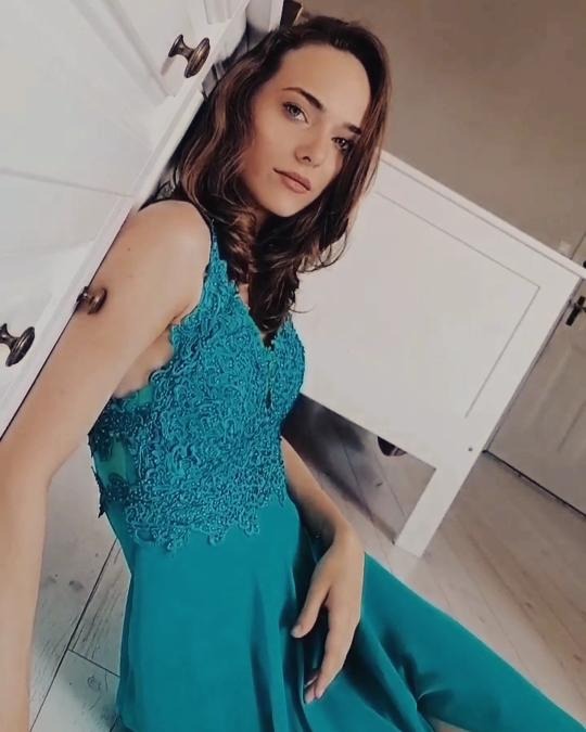 Portret kobiety w turkusowej sukni z koronkową górą, opierającej się o białą szafkę z mosiężnymi uchwytami, na podłodze z jasnego drewna.