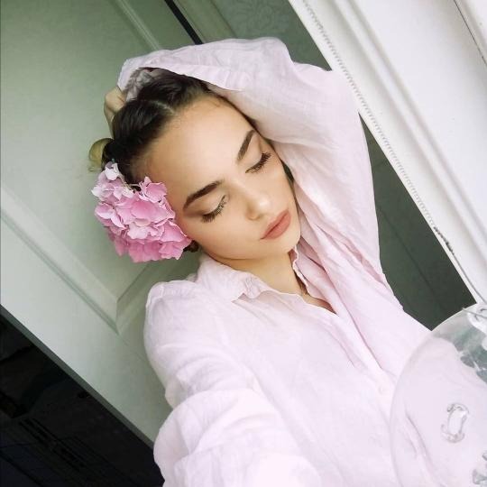 Portret kobiety z zamkniętymi oczami, różowym kwiatem hortensji we włosach i w jasnoróżowej koszuli, oparta o białą ramę okna, delikatne światło.