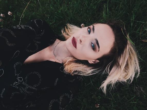 Kobieta z blond pasemkami i wyrazistym makijażem leży na trawie w czarnej sukience z subtelnym wzorem.