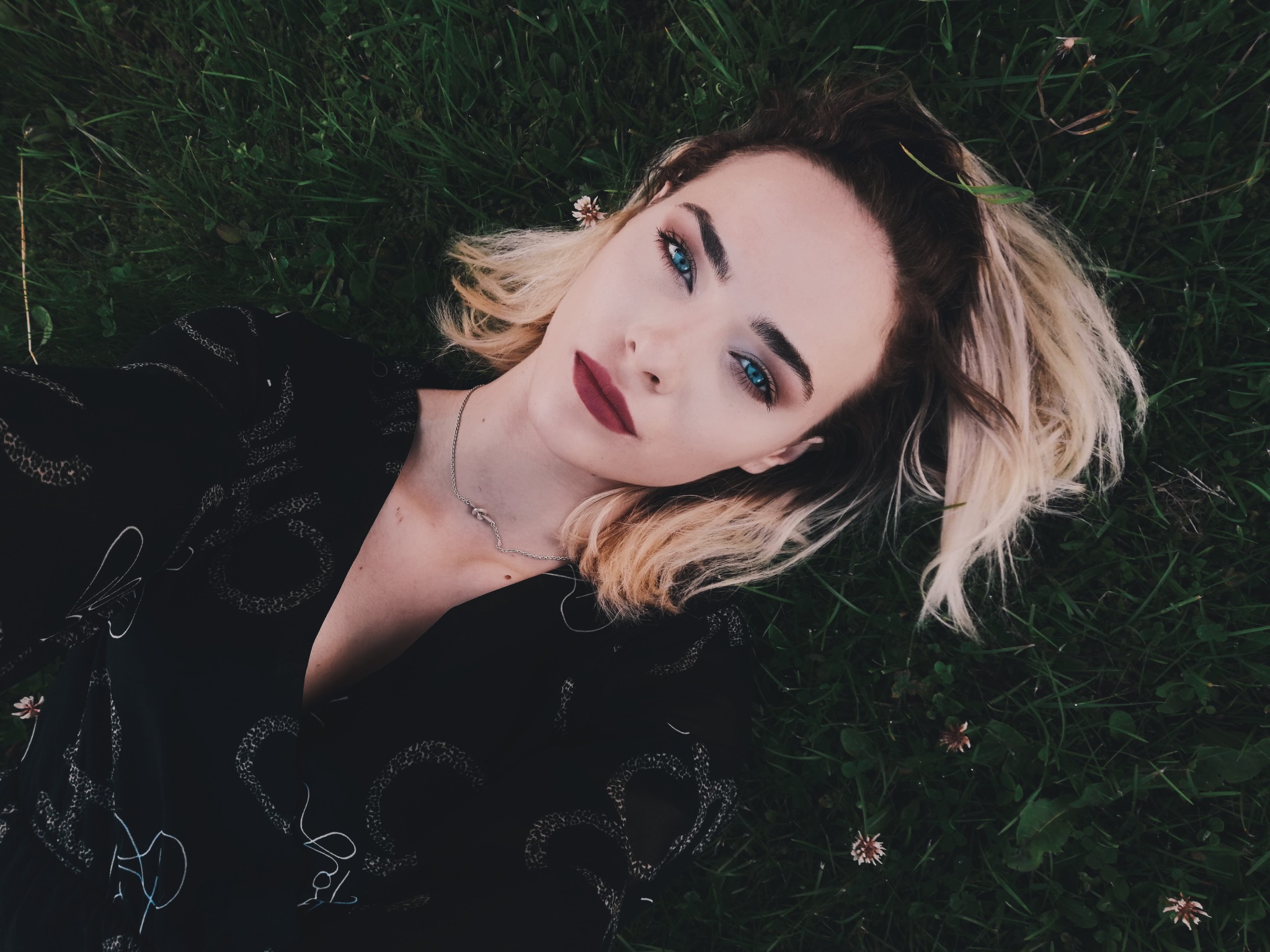 Portret kobiety z niebieskimi oczami leżącej na trawie w Szczecinie, ubrana w czarną sukienkę w jasne wzory, z delikatnym srebrnym naszyjnikiem.