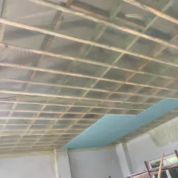 Montaż płyty gipsowo-kartonowej na drewnianej konstrukcji sufitu, zabezpieczonej folią; widoczne rusztowanie i fragment ściany w surowym stanie.