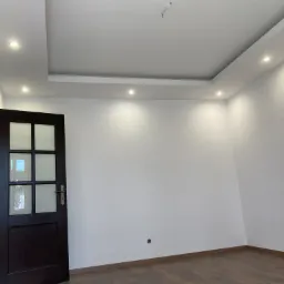 Wnętrze pokoju po remoncie: białe ściany, drewniana podłoga, ciemne drzwi z szybami i oświetlenie sufitowe punktowe.