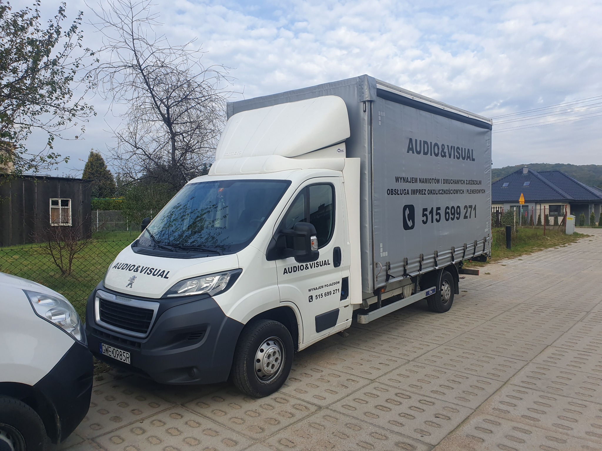 Biały samochód dostawczy z szarą plandeką, z napisem 'AUDIO&VISUAL' oraz numerem telefonu, zaparkowany na brukowanej drodze. Widoczny fragment drugiego samochodu.