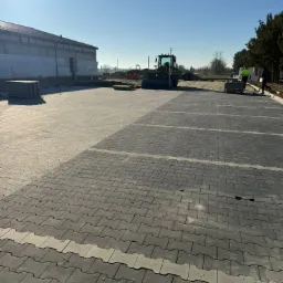 Rozległy parking z szarej kostki brukowej, wykończony jaśniejszymi pasami. W tle budynek w budowie i sprzęt budowlany, a także pracownicy w odblaskowych kamizelkach.