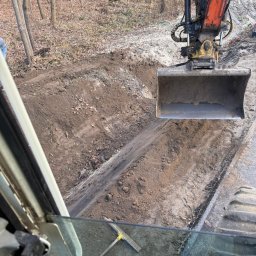 GARDEN MAX MARCIN KOWAL - Koparka w trakcie prac ziemnych, widok z kabiny operatora. Wykopany rów w ziemi, w tle las. Przygotowanie terenu pod utwardzenie drogi.