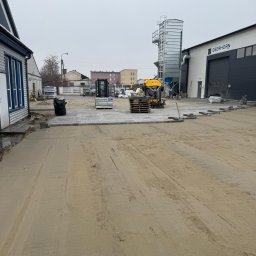 GARDEN MAX MARCIN KOWAL - Prace brukarskie w toku: mini koparka, palety z kostką i fragment ułożonej nawierzchni na placu budowy. W tle budynki i elementy infrastruktury.