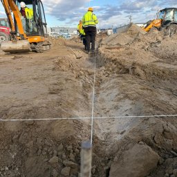 GARDEN MAX MARCIN KOWAL - Prace ziemne: Mini koparka, niwelator i ekipa w odblaskowych kamizelkach przy wytyczaniu terenu pod budowę. Widoczne liny wyznaczające osie.