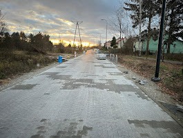 Układanie kostki brukowej na drodze osiedlowej, widoczne fragmenty ułożonej nawierzchni, palety z kostką i słup oświetleniowy. Zachód słońca w tle.