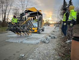 Żółty układacz kostki brukowej w trakcie pracy na drodze; widoczni pracownicy w odblaskowych kamizelkach i kostka brukowa w różnych etapach ułożenia.