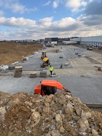 Rozległy plac budowy z częściowo ułożoną szarą kostką brukową, maszyny budowlane i stosy kostki w tle, przy pochmurnym niebie. Widok z góry.