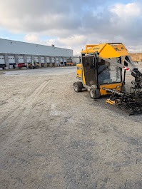 Żółta maszyna do układania kostki brukowej na placu budowy; w tle budynek magazynowy z rampami załadunkowymi i pochmurne niebo.