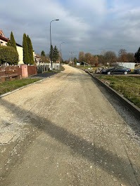 Utwardzona droga gruntowa w trakcie prac, widok z dołu. Po prawej krawężnik, w tle zabudowa jednorodzinna i pochmurne niebo. Perspektywa zbieżna.