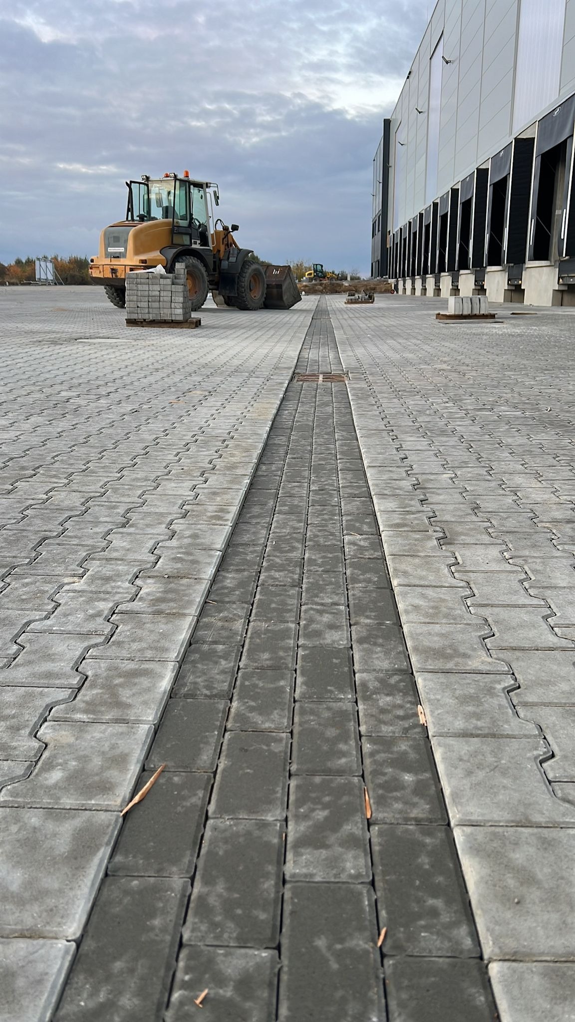 Ułożona szara kostka brukowa z centralnym pasem z ciemniejszej kostki. W tle budynek magazynowy i maszyny budowlane. Całość w pochmurny dzień.