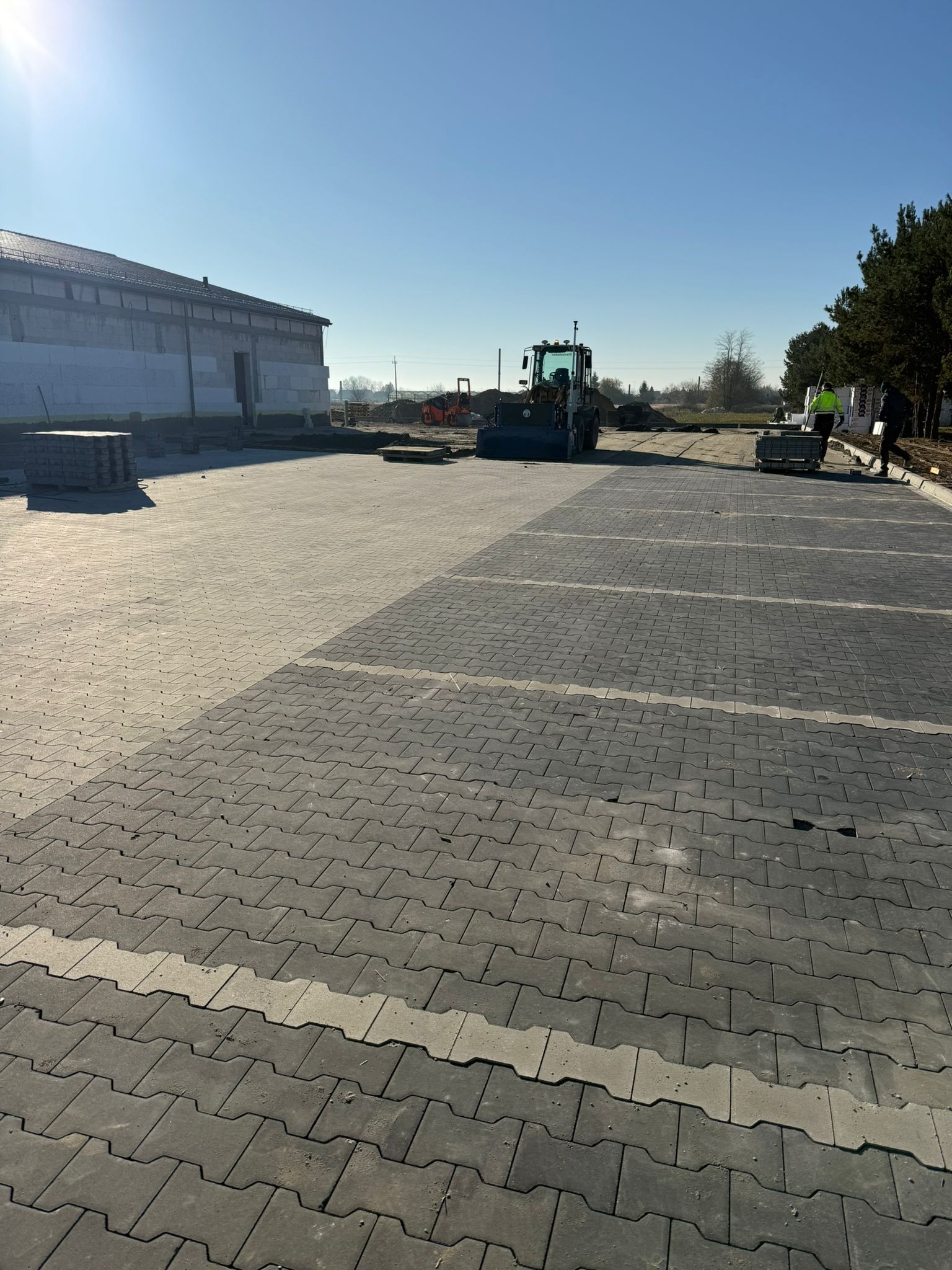 Rozległy parking z szarej kostki brukowej, wykończony jaśniejszymi pasami. W tle budynek w budowie i sprzęt budowlany, a także pracownicy w odblaskowych kamizelkach.