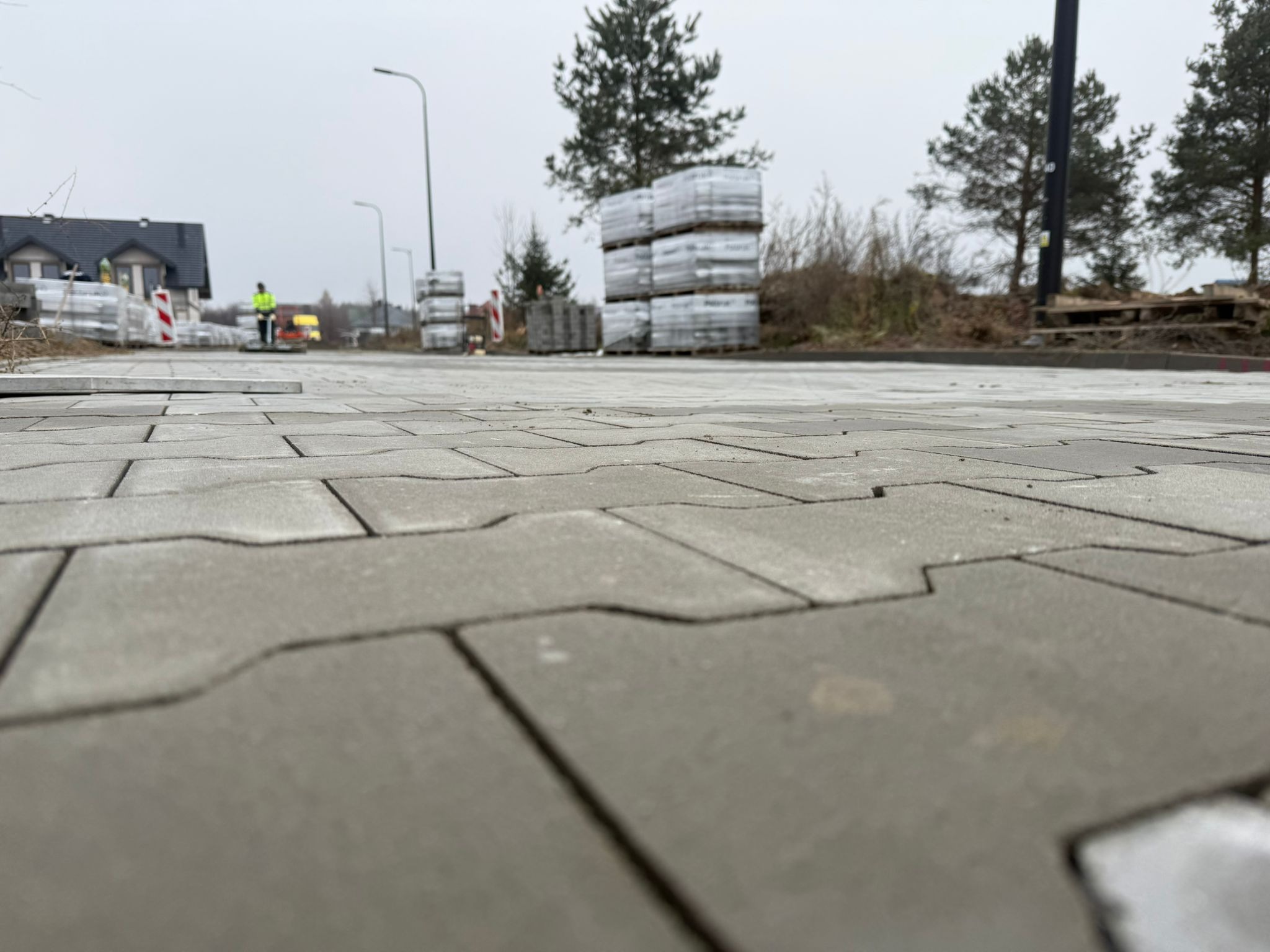 Ułożona szara kostka brukowa z paletami kostki w tle, widok z niskiej perspektywy. W oddali pracownik w odblaskowej kamizelce i fragment domu.